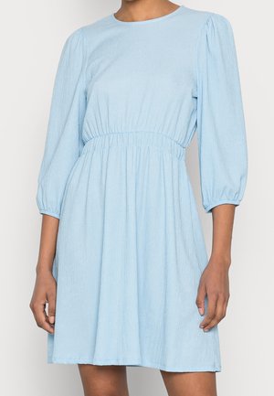Femme portant une robe bleu clair jusqu'aux genoux, avec des manches longues bouffantes et une taille élastique, debout devant un fond uni.