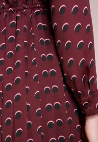 Abito in tessuto bordeaux con motivo circolare nero e rosa, design plissettato e dettaglio con bottoni sui polsini delle maniche. Materiale morbido e testurizzato.