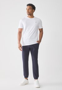 Man standing wearing white short-sleeve t-shirt with emblem, navy jogger pants, and white sneakers against a plain light background.
