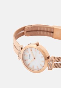 Rožnato zlata mrežasta zapestnica ure z belo številčnico, rožnato zlatimi oznakami za ure in kristalnimi detajli na zatičih. Ima okroglo ohišje in krono.