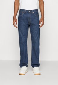 Dark blue denim jeans with straight cut, five pockets, contrasting stitching, and metal button closure; worn with white sneakers.