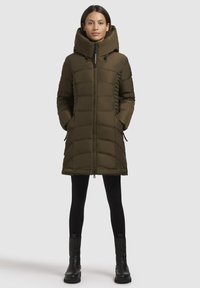 Vrouw in een olijfgroen gewatteerd winterjack tot op de knie met hoge kraag, zwarte leggings en zwarte enkellaarsjes, rechtopstaande.