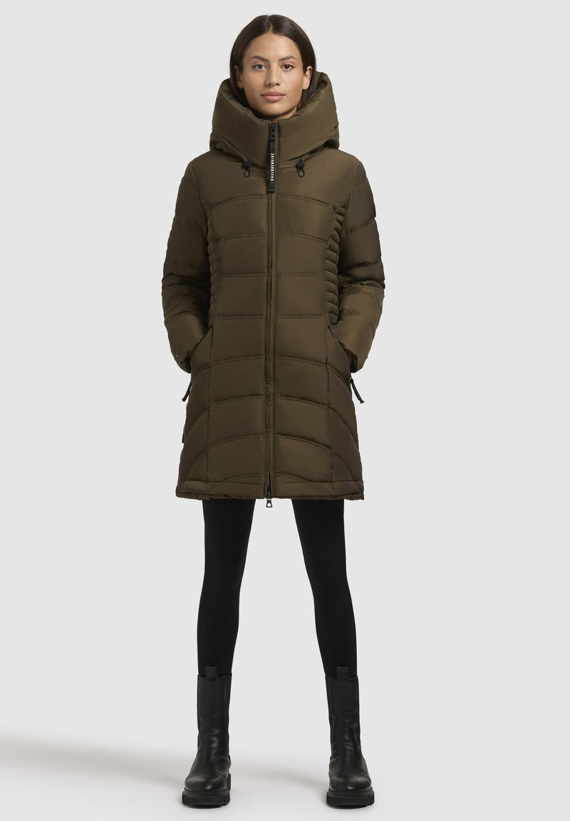 Frau in einem knielangen olivgrünen, gepolsterten Wintermantel mit hohem Kragen, schwarzen Leggings und schwarzen Stiefeletten, die aufrecht steht.
