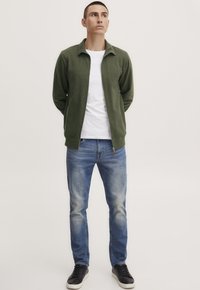 Grønn zip-up jakke over en hvit T-skjorte, kombinert med blå denimjeans og svarte joggesko. Glatt tekstur og avslappet passform.