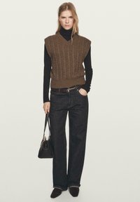 Braune Strickweste mit Kabelmuster über einem schwarzen Rollkragenpullover, kombiniert mit dunklen Jeans und schwarzen Accessoires. Verfügt über weite Ärmel und einen V-Ausschnitt.