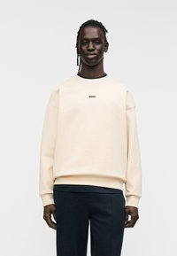 Sweat-shirt beige avec un col rond, arborant le logo "BOSS" en noir. Coupe oversized avec poignets et ourlet côtelés. Associé à un pantalon foncé.