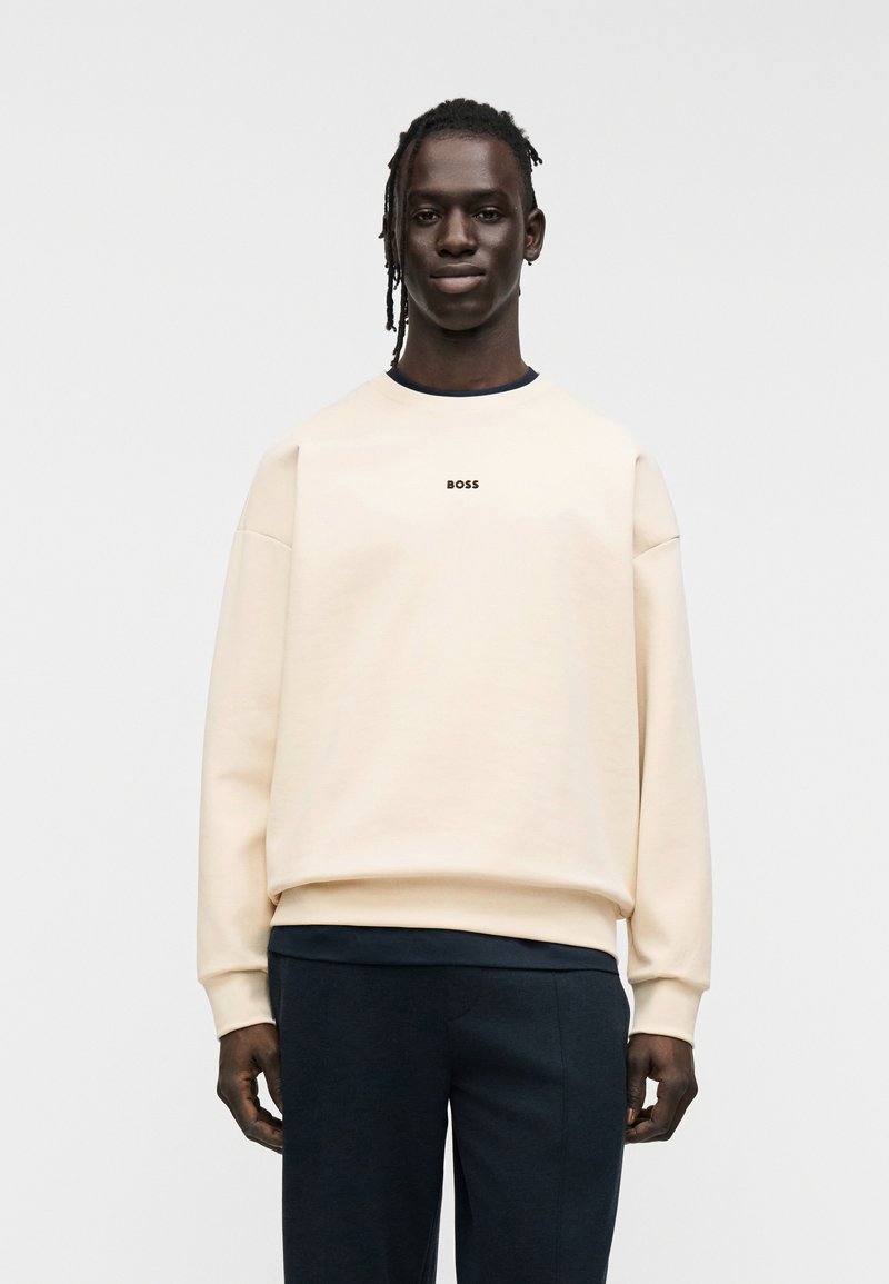 Sweat-shirt beige avec un col rond, arborant le logo "BOSS" en noir. Coupe oversized avec poignets et ourlet côtelés. Associé à un pantalon foncé.