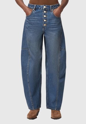 Person iført blå jeans med høj talje og vide ben med knaplukning foran og hænder i lommerne, kombineret med brune ankelstøvler med hæl.