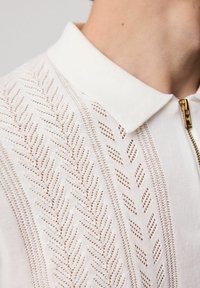 Polo de punto blanco con un intrincado diseño de hojas. Presenta un cuello con solapa, tejido texturizado y una cremallera dorada.