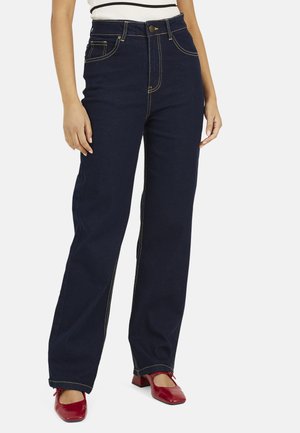 Vrouw draagt donkerblauwe jeans met hoge taille en wijde pijpen en rode lage hakken met vierkante neus, staand met ontspannen handen langs het lichaam.