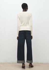 Maglione bianco a costine con orlo smerlato, abbinato a jeans a gamba larga blu scuro con risvolti, e scarpe con tacco marroni.