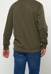 Oliivinvihreä huppari pitkillä hihoilla ja ribbireunuksilla, yhdistettynä sinisiin farkkuihin, joissa on takataskut kaksinkertaisella ompelulla.