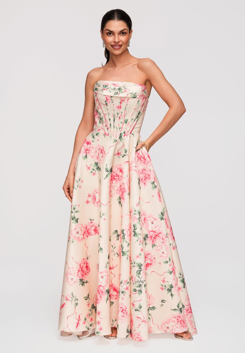 Femme portant une robe ivoire longue sans bretelles avec un imprimé floral rose, posant avec une main dans la poche et souriante.