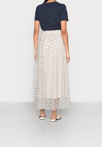Lange doorschijnende rok met polkadots in crème en marineblauwe stippen, gecombineerd met een donkerblauw T-shirt met korte mouwen. Platte, lichtgekleurde schoenen.