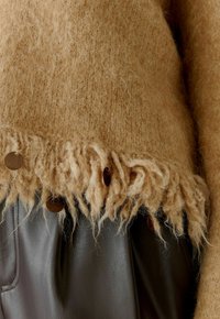 Flauschiger beiger Pullover mit fransigem Saum, ausgestattet mit braunen runden Knöpfen, kombiniert mit dunklen Lederhosen, die Textur und Kontrast betonen.