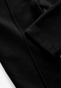 Detailní záběr na černý pletený oděv, který ukazuje strukturovanou tkaninu, viditelné švy a ohrnuté manžety, zdůrazňující materiál a tvar.