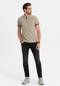 Beige polo skjorta med svart kant, kortärm, och treknappsstängning; kombinerad med smala svarta jeans och ljusa sneakers.