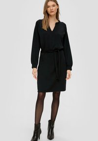 Czarna sukienka z dekoltem w kształcie litery V i marszczonym wcięciem w talii, z długimi rękawami i paskiem do przewiązania. Noszona z czarnymi botkami sięgającymi kostki i przezroczystymi rajstopami.