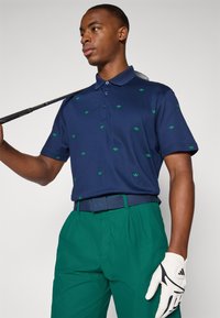 Polo marino con pequeños patrones de hojas verdes, con una tapeta de tres botones, combinado con pantalones verdes, belt negro texturizado, y guante de golf blanco.