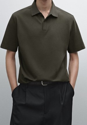 Homme portant un polo à manches courtes vert olive rentré dans un pantalon noir avec une ceinture noire, mains dans les poches, sur fond uni.