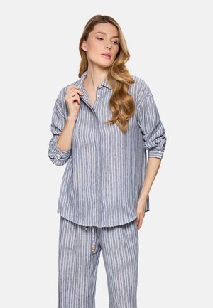 Mujer con cabello largo y ondulado, vestida con camisa de botones a rayas verticales azules y blancas y pantalones a juego, posando con una mano cerca del cuello.