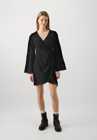 Μαύρο φόρεμα περιτυλίγματος από υφασμάτινη υφή με V-λαιμόκοψη, μακριά μανίκια καμπάνα και ασύμμετρο στρίφωμα. Φοριέται με μαύρες μπότες μέχρι τον αστράγαλο και λευκές κάλτσες.