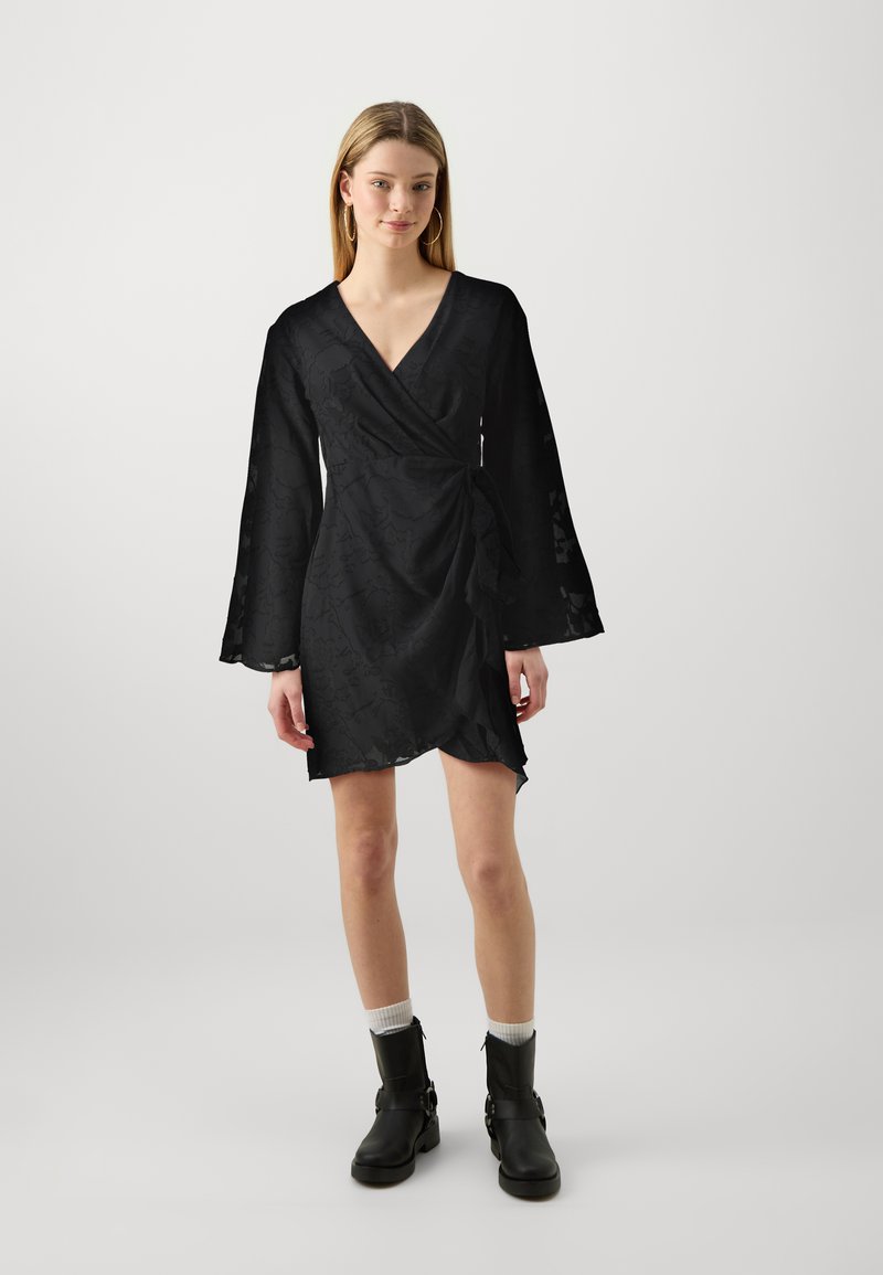 Μαύρο φόρεμα περιτυλίγματος από υφασμάτινη υφή με V-λαιμόκοψη, μακριά μανίκια καμπάνα και ασύμμετρο στρίφωμα. Φοριέται με μαύρες μπότες μέχρι τον αστράγαλο και λευκές κάλτσες.