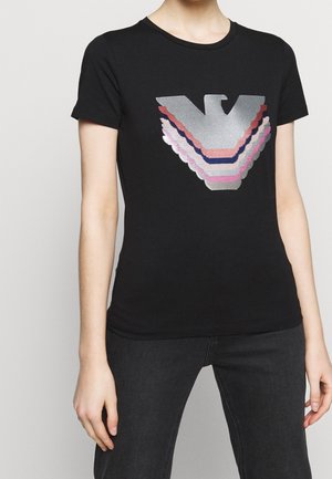 Personne portant un t-shirt noir à manches courtes avec un grand motif géométrique d'aigle argenté, rose et bleu marine sur le devant, associé à un jean foncé.