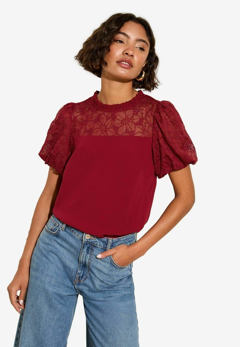 Blusa bordeaux con sovrapposizione di pizzo floreale sulle spalle, maniche a sbuffo e scollatura tonda, abbinata a jeans blu a vita alta.