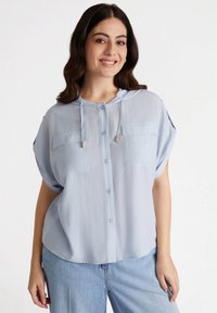 Camicia a maniche corte azzurro chiaro con cappuccio, dotata di due tasche sul petto e chiusura frontale con bottoni. Realizzata in un tessuto trasparente e texture.