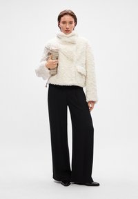 Λευκή φούξια κοντή ζακέτα με ψηλό γιακά, συνδυασμένη με μαύρους παντελόνες φαρδιάς γραμμής. Η μοντέλα κρατάει μια μπεζ υφασμάτινη τσάντα.