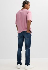 Camiseta rosa de manga corta, de corte relajado, combinada con unos vaqueros azul oscuro y zapatillas blancas. El modelo está de pie con las manos en los bolsillos, dando la espalda.