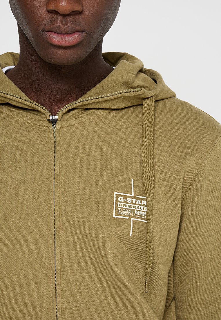 Olivegrüne Zip-Hoodie aus strukturiertem Stoff, mit weißem, gesticktem Logo auf der Brust und Kordelzug an der Kapuze.