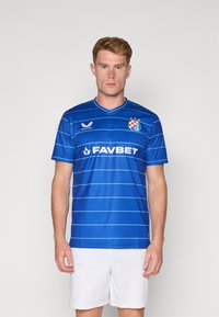 Maglia da calcio blu con strisce orizzontali, colletto a girocollo e loghi sul petto. Indossata con pantaloni corti bianchi da atleta.