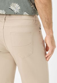 Pantaloni beige con una texture liscia, dotati di una singola tasca posteriore e di un discreto logo vicino alla vita. Il design è semplice e funzionale.