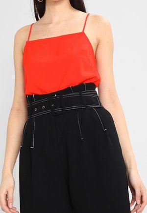 Vrouw draagt een mouwloze rode top met vierkante hals, ingestopt in een zwarte broek met hoge taille, brede riem en witte stiksel details.