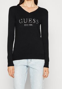 Zwarte lange mouwen trui met V-hals, met zilveren rhinestone merknaam "GUESS EST. 1981" aan de voorkant, geribde manchetten en zoom.