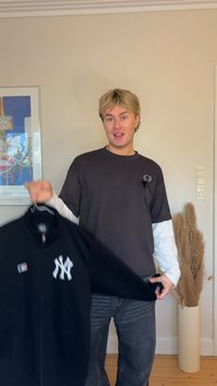 Young person with short blond hair holds a black New York Yankees jacket, standing in a room with a framed poster and dried plants.