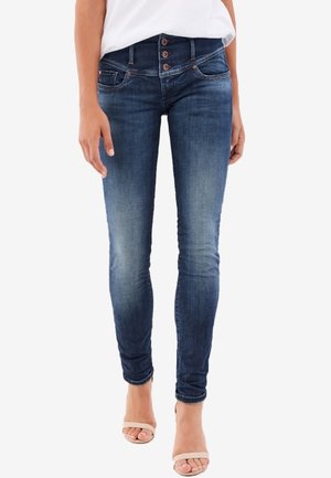 Mujer vistiendo jeans ajustados azul oscuro con cierre de botones de tiro alto, combinados con tacones abiertos beige y una blusa blanca.