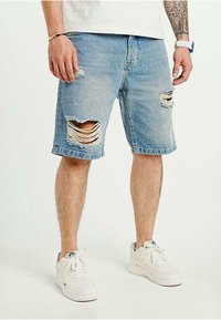 Ljusblå denimshorts med fransad nederkant och flera slitna lappar. Kombinerat med vita sneakers. Tätt sittande silhuett.