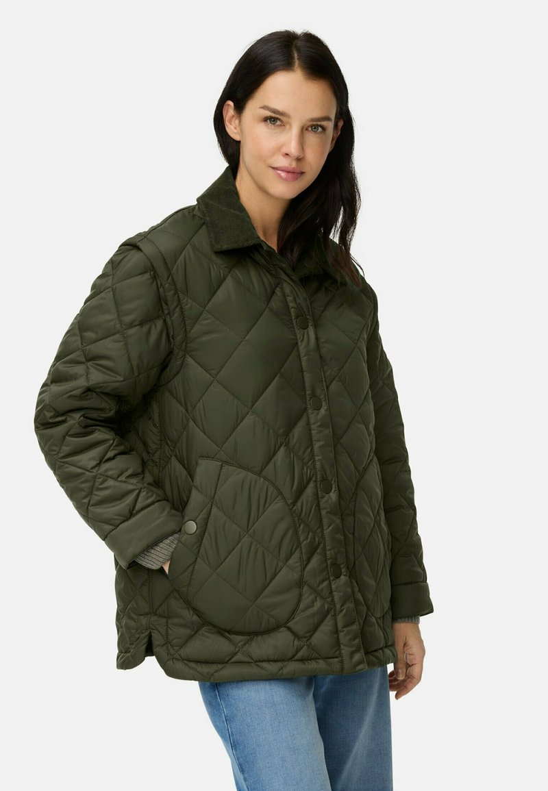 Vrouw in een olijfgroene gewatteerde jas met grote zakken en knoopsluiting, gecombineerd met lichtblauwe jeans, staande tegen een effen achtergrond.