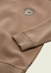 Sweatshirt marron avec un graphique d'ours et le texte "JOURS SYMPAS". Poignets et ourlet côtelés, texture douce, et une petite étiquette de logo sur le côté.