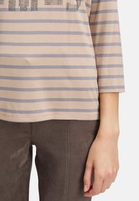 Beige- und graustreifiges Oberteil mit Dreiviertelärmeln, das mit glänzenden Verzierungen ausgestattet ist. Kombiniert mit dunkelbraunen Wildlederhosen.