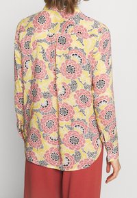 Blusa floral en amarillo, rosa y negro con un estampado de grandes flores y hojas; mangas largas y detalle de dobladillo dividido.