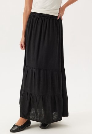 Woman wearing a long black tiered skirt with an elastic waistband and black ballet flats, standing with one hand on her hip.