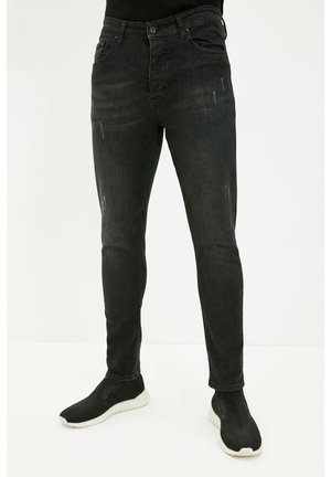 Person wearing slim-fit black jeans with subtle distressed details and black slip-on sneakers with white soles.