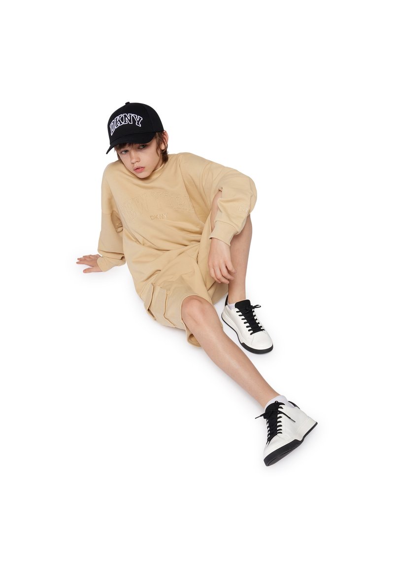 Robe-sweatshirt oversize beige avec le texte en relief "NEW YORK DKNY", associée à une casquette noire et des baskets montantes noir et blanc.
