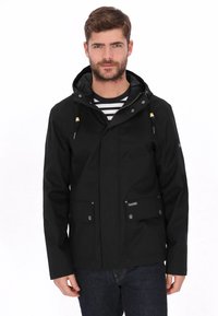 Zwarte waterdichte jas met capuchon, twee voorzakken, drukknopen en verstelbare trekkoorden. Draagt over een gestreept shirt.