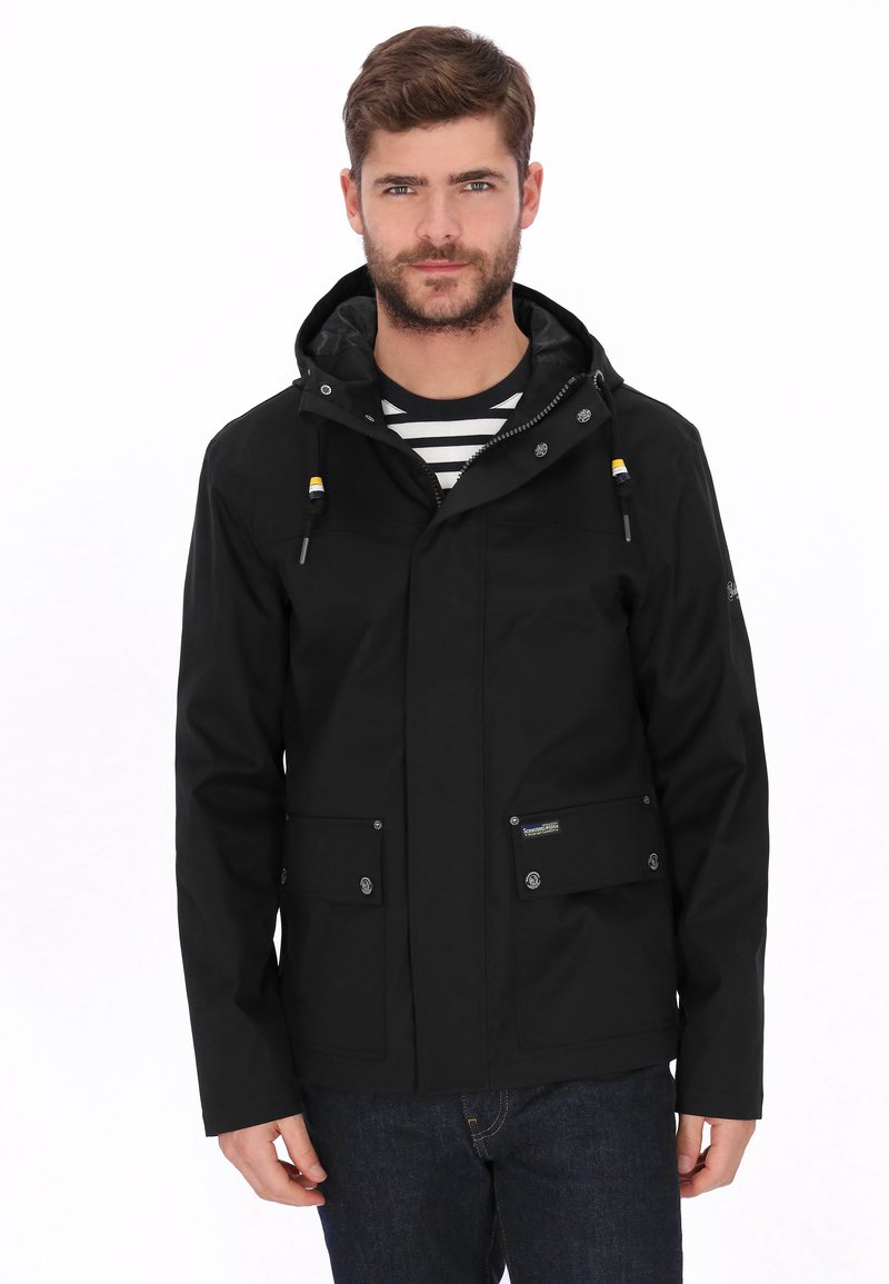 Zwarte waterdichte jas met capuchon, twee voorzakken, drukknopen en verstelbare trekkoorden. Draagt over een gestreept shirt.