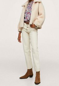 Persona que lleva una chaqueta de borreguillo color crema, blusa floral, pantalones crema de tiro alto con botón y botas de ante marrón hasta el tobillo.